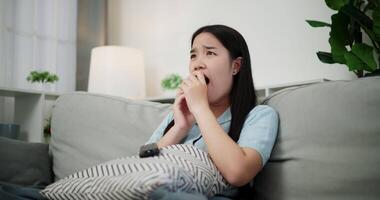 Handheld shot, Young Asian woman watching an exciting movie with a shocked and thrilled expression while sitting on a sofa in her living room at home. Leisure activity, Lifestyle concept. video