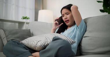 Handheld low angle view shot, An upset young woman is talking on the phone to someone with an angry and irritated look on her face while sitting on the sofa in living room at home. video