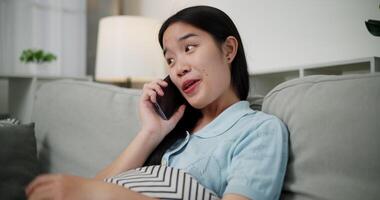 Handheld low angle view shot, Cheerful young woman is having fun talking on smartphone with her friend, smiling and laughing happily while sitting on the sofa in the living room at home. video