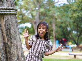 Cute little girl is peeking behind a tree, having fun playing playing peek-a-boo in a sunny garden. Smiling child in the park photo