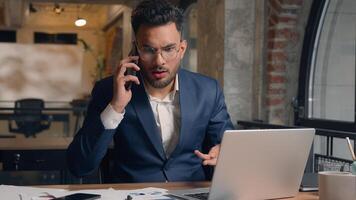Stressed multitasking businessman Indian Arabian male guy annoyed employer manager consultant talking mobile phone shout working laptop writing documents anxious business man problem overwork office photo