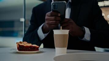 Unrecognizable man African businessman entrepreneur with mobile phone browsing chatting make picture photo blogger vlogger male at table with smartphone in cafe with cake coffee messaging in cafeteria