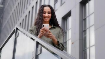 Smiling Caucasian Hispanic happy satisfied woman user lady standing un city outdoors on stairs chatting texting searching in net with mobile app phone browsing smartphone friendly communication online photo