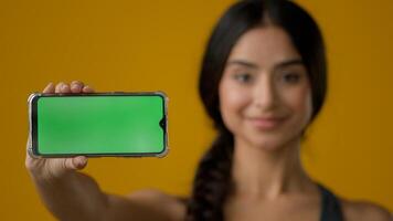 Close up Indian ethnic smiling woman female girl in yellow studio holding phone horizontal showing green empty mobile screen of smartphone chroma key display for advertisement online add offer in cell photo