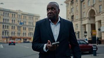 Ethnic adult man African middle-aged businessman entrepreneur with phone in city booking taxi with mobile app waiting car confused upset looking around problem with navigation mistake navigator wait photo