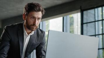 Serious 40s bearded businessman entrepreneur employee standing in office looking at papers report information papers check data in online net service in computer typing working paperwork deadline photo