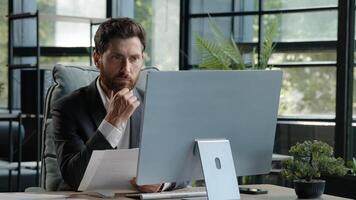 Caucasian pensive bearded man boss manager adult 40s businessman sitting in office busy with paperwork and computer check data in papers search information look at screen corporate company app project photo