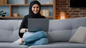 Muslim Islamic woman in hijab sitting at home living room relaxing on sofa couch using laptop online website ordering booking in internet shop working studying remote distant chatting typing computer photo