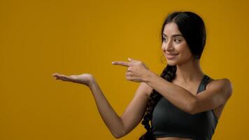 Portrait in studio Indian 20s woman posing on yellow background pointing to side at palm arm hand point advertisement aside recommend recommendation prize sign girl lady showing to empty copy space photo