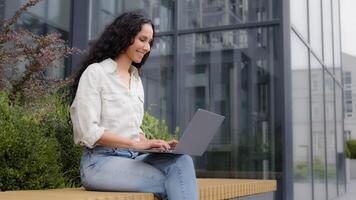 Smiling businesswoman sits on bench near office building on terrace with laptop chatting with clients colleagues works writes article. Girl student e-learning with wireless computer using free Wi-Fi photo