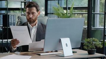 Serious adult 40s bearded pensive thoughtful businessman working at computer in office with paperwork. Professional employee worker typing thinking over online project check data in documents contract photo