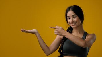 Portrait in studio Indian 20s woman posing on yellow background pointing to side at palm arm hand point aside recommend recommendation sign girl lady showing to empty copy space looking at camera photo