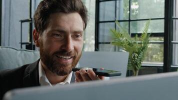 Happy adult 40s male manager bearded businessman sitting at workplace with computer smiling recording audio message sending voicemail using assistant software application on smartphone mobile app chat photo
