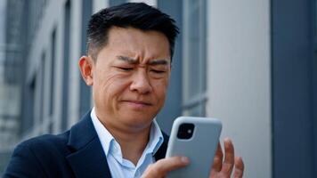Close up angry worried stressed Asian middle-aged 40s man user businessman employer entrepreneur trying browsing on broken cellphone outdoors in city annoyed with mobile app error connection problem photo