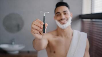 Funny hispanic indian arab naked man 30s bearded guy standing in bathroom at home with white soapy foam on beard shaving gel smiling showing razor looking at camera getting ready to shave facial care photo