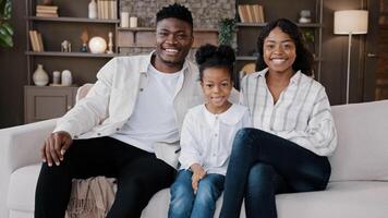 African family sit down on comfortable couch in living room. Biracial parents and child smiling looking at camera. Cheerful daughter with mum and dad at home sitting on sofa in new house, real estate photo