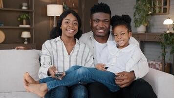 Carefree african american father, mather relaxing with biracial daughter child girl at hme holidays sitting on sofa at home watching television together. Cheerful family relaxing watching tv movie photo