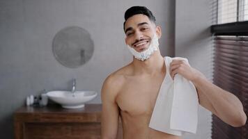 Close up male portrait in bathroom hispanic indian arabian naked man bearded guy posing smiling with white soapy foam on beard shaving gel holding bath towel looking at camera getting ready to shave photo