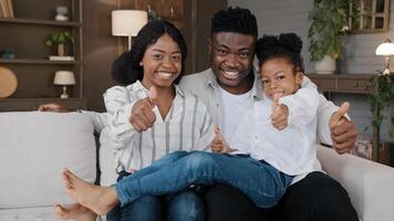 Happy African family young guardian parents and little daughter adopted girl child sitting on sofa at home own apartment new house looking at camera show thumbs up approve recommend positive attitude photo