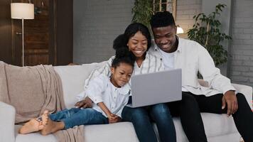 Happy African family and cute kid child girl daughter using laptop looking at computer screen enjoying watching funny social media having online call conference relaxing on sofa at home photo