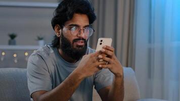 Serious busy Arabian Indian man bearded male in glasses typing browsing in smartphone texting chatting using mobile app messaging at home sitting on couch at night playing game e-commerce shopping photo