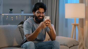 Smiling Arabian Indian man bearded male looking in smartphone smile texting with friend girlfriend messaging chatting at home sitting on couch at night playing game in mobile app watching funny photo