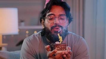 Portrait arabian happy indian male face bearded man wears festive birthday pink hat and glasses holds cupcake makes wish joyful congratulating at home blowing burning candles on cake celebrating party photo