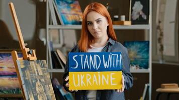 Red-haired talented artist stands posing in art studio workshop against background of drawing picture, painter holds cardboard banner stand with Ukraine support freedom of Ukrainians anti war poster photo