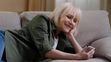 Happy relaxed Caucasian old senior mature woman holding smart phone looking at cellphone laughing lying on sofa couch enjoying using mobile apps for shopping having fun watching funny in media photo