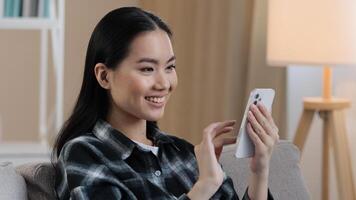 Smiling happy Asian woman typing swipe touchscreen of phone Korean girl cheerful choosing shopping in online e-commerce app female enjoy mobile website play game at home follow post in social media photo