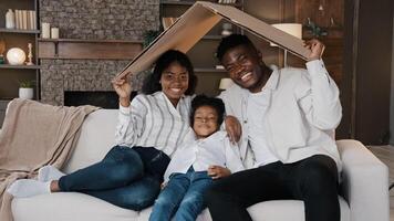 African couple sitting on couch under cardboard roof smiling looking at camera active happy daughter girl running sits on sofa. Relocation day to new home moving to own apartment homeowner portrait photo