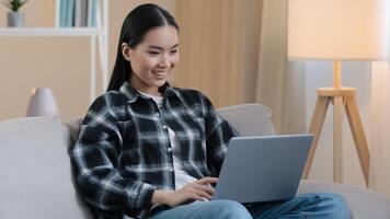 Smiling asian woman happy student girl freelancer sitting on couch using laptop typing message email lady chatting on computer browsing internet media at home easy e-learning studying working online photo