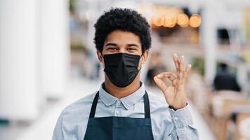 Masked african american male cafe worker waiter salesman in apron medical face mask successful man small business owner looking camera shows ok okay agree fine good sign great gesture pandemic rules photo