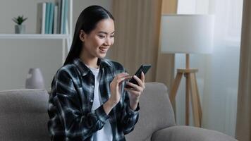 Asian happy smiling girl woman sitting home sofa looking at mobile phone chatting browsing using wifi web service watching online app texting with friends smartphone ordering internet shopping photo