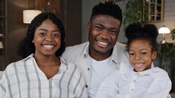 Happy African family portrait. Parents with cheerful daughter girl looking at camera laughing cuddling at home smiling toothy. Positive mother, father and child hugging on sofa. Dental health concept photo