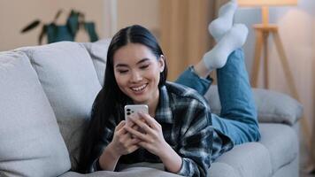 Asian female relaxing girl woman lying on couch at home enjoy chat typing app mobile phone relax watch viral live play game follow like post in social media share and send smile emoji scroll web photo