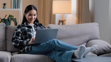 Smiling asian woman using laptop casual browsing searching online play computer game. Korean student girl freelancer sitting on couch e-learning internet education chatting studying working with wi-fi photo