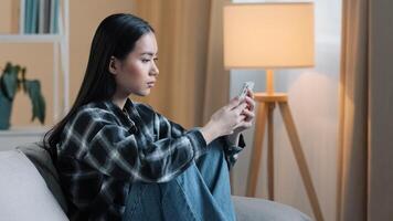 Side view of sad bored Asian woman girl sitting on sofa at home with telephone serious unhappy lady scrolling browsing in smartphone and using mobile phone app watching social media news feed typing photo