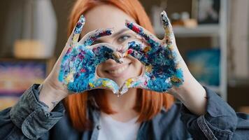 Close up head shot red-haired 20s smiling girl in eyeglasses smiling toothy friendly with love painter woman looking at camera holding dirty in paints hands in heart shape shows gesture of kindness photo
