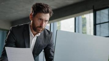 Serious 40s bearded businessman entrepreneur employee standing in office looking at papers report has problem with papers check data in online net service in computer typing working paperwork deadline photo