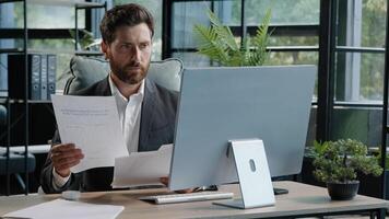Busy adult 40s bearded pensive thoughtful businessman working at computer in office with paperwork. Professional employee worker typing thinking over online project check data in documents contract photo