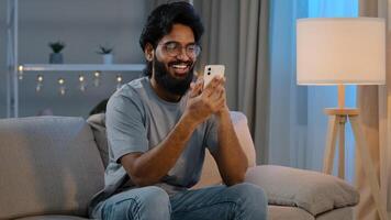 Happy arabian indian male millennial man holding modern smartphone texting message at home sitting on couch in evening bearded guy in glasses using mobile app watching funny online laughing photo