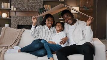 African couple sitting on couch under cardboard roof smiling looking at camera active happy daughter girl running sits on sofa. Relocation day to new home moving to own apartment homeowner portrait photo