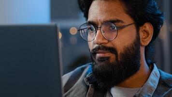 Close-up bearded male face reflection light from laptop monitor in glasses. Busy serious focused Arab Indian man looking at computer working freelance in evening playing game at night surfing in net photo