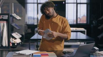 Arabian Indian male business man working busy interior designer talking with client online discuss on mobile phone fabric tissue samples write in notepad textile order in workshop modern design studio photo