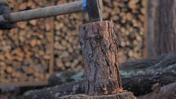 An axe strikes a solid log resting on a sturdy stump, sending splinters flying. In the background, stacked firewood creates a cozy outdoor atmosphere. video