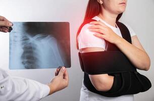 The doctor holds in his hand a medical x-ray of a dislocated humerus and a fractured collarbone against the background of a girl patient whose shoulder hurts. Fixing bandage for the shoulder joint. photo