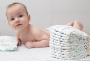 A little boy at the age of 5 months lies in a diaper on a white background and looks at the camera. Stack of diapers, anti-leak, soft and comfortable. Copy space for text photo