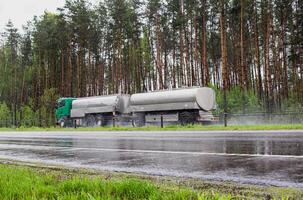 A truck with a tank semi-trailer transports liquid cargo on a slippery wet road in summer against the backdrop of a forest. Transportation of goods in bad weather. Sanctions photo