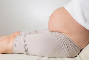 A pregnant girl with a big belly sits against a white wall. Concept of testing amniotic fluid to check for genetic diseases in the fetus, amniocentesis. Copy space for text, preparation for childbirth photo
